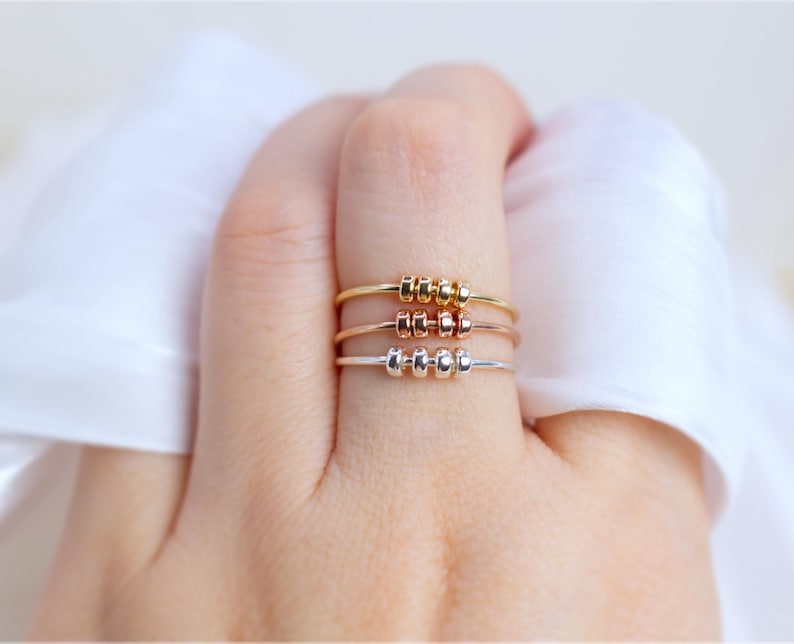 May include: Three delicate rings in gold, rose gold, and silver, each featuring three small, evenly spaced beads. The rings are displayed on a hand, showcasing their minimalist design and varying metallic finishes. The background is a soft, white fabric.