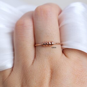 May include: Rose gold ring with three small, cylindrical beads centered on the band. The ring is displayed on a person's finger, with a white fabric in the background. The word "coco" is printed on the ring.