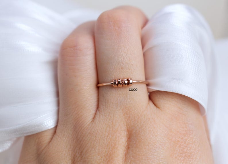 May include: Rose gold ring with three small, cylindrical beads centered on the band. The ring is displayed on a person's finger, with a white fabric in the background. The word "coco" is printed on the ring.