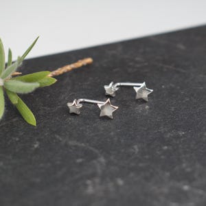 Puede incluir: Dos pendientes de color plateado, cada uno con una estrella peque&ntilde;a y una estrella m&aacute;s grande conectadas por una barra de metal curva. Las piezas de joyer&iacute;a tienen un acabado met&aacute;lico pulido y siguen la curva del l&oacute;bulo de la oreja.