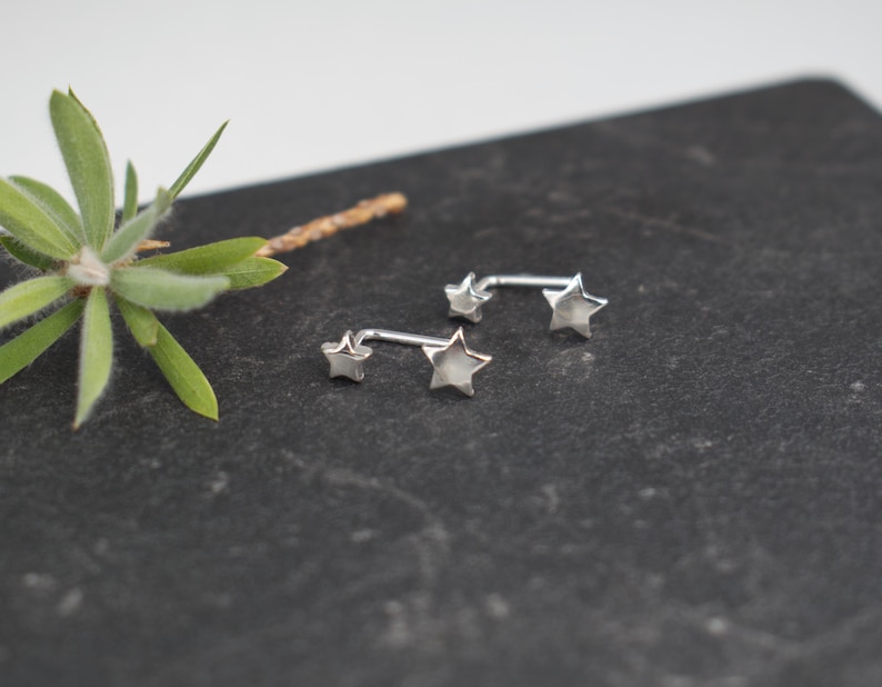 Puede incluir: Dos pendientes de color plateado, cada uno con una estrella peque&ntilde;a y una estrella m&aacute;s grande conectadas por una barra de metal curva. Las piezas de joyer&iacute;a tienen un acabado met&aacute;lico pulido y siguen la curva del l&oacute;bulo de la oreja.