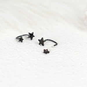 Puede incluir: Un conjunto de piezas de joyer&iacute;a de metal negro con dise&ntilde;os en forma de estrella. El set incluye dos pendientes o anillos abiertos y curvados con estrellas en los extremos, y una peque&ntilde;a pieza de pendiente tipo bot&oacute;n con forma de estrella.