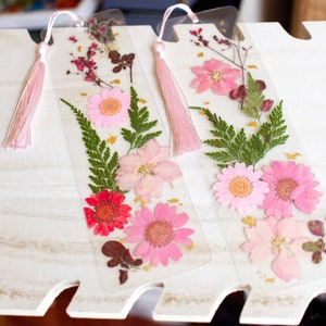 May include: Two clear bookmarks with pink tassels. Each bookmark features pressed flowers and greenery, including pink and red flowers, green ferns, and gold accents.