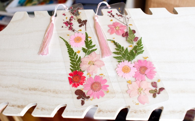 May include: Two clear bookmarks with pink tassels. Each bookmark features pressed flowers and greenery, including pink and red flowers, green ferns, and gold accents.