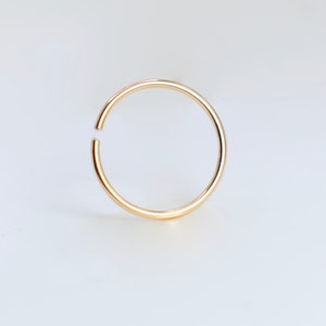 May include: A thin, circular gold hoop ring with a smooth, polished finish. The ring features a small, distinct gap along its circumference, creating an open-ended design that allows for flexibility and ease of use in various piercing placements.