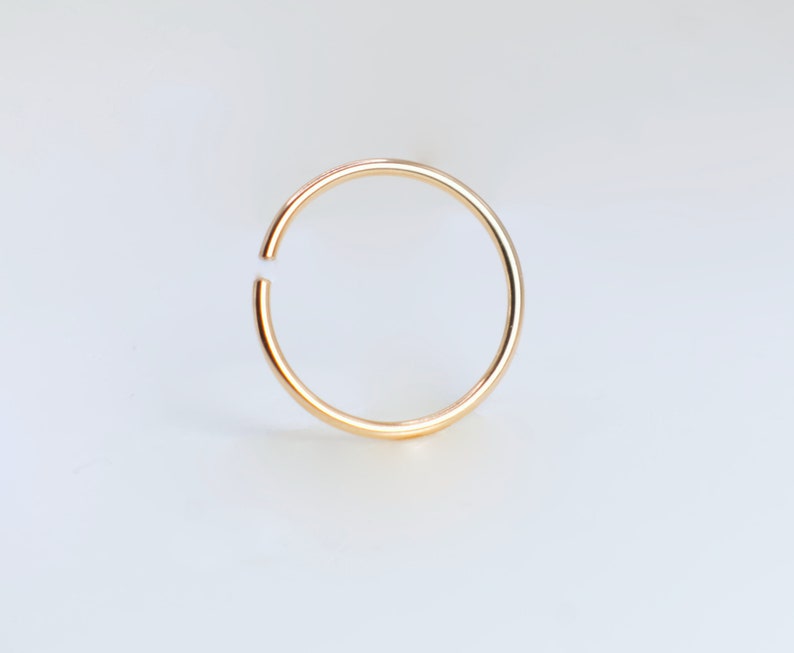 May include: A thin, circular gold hoop ring with a smooth, polished finish. The ring features a small, distinct gap along its circumference, creating an open-ended design that allows for flexibility and ease of use in various piercing placements.