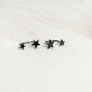 Puede incluir: Dos anillos abiertos de metal negro, cada uno con dos detalles en forma de estrella de diferentes tama&ntilde;os. Los anillos tienen un acabado oscuro y liso, con un dise&ntilde;o de banda curva que permanece abierta en la parte superior, mostrando una estrella en cada extremo.