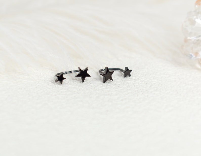 Puede incluir: Dos anillos abiertos de metal negro, cada uno con dos detalles en forma de estrella de diferentes tama&ntilde;os. Los anillos tienen un acabado oscuro y liso, con un dise&ntilde;o de banda curva que permanece abierta en la parte superior, mostrando una estrella en cada extremo.