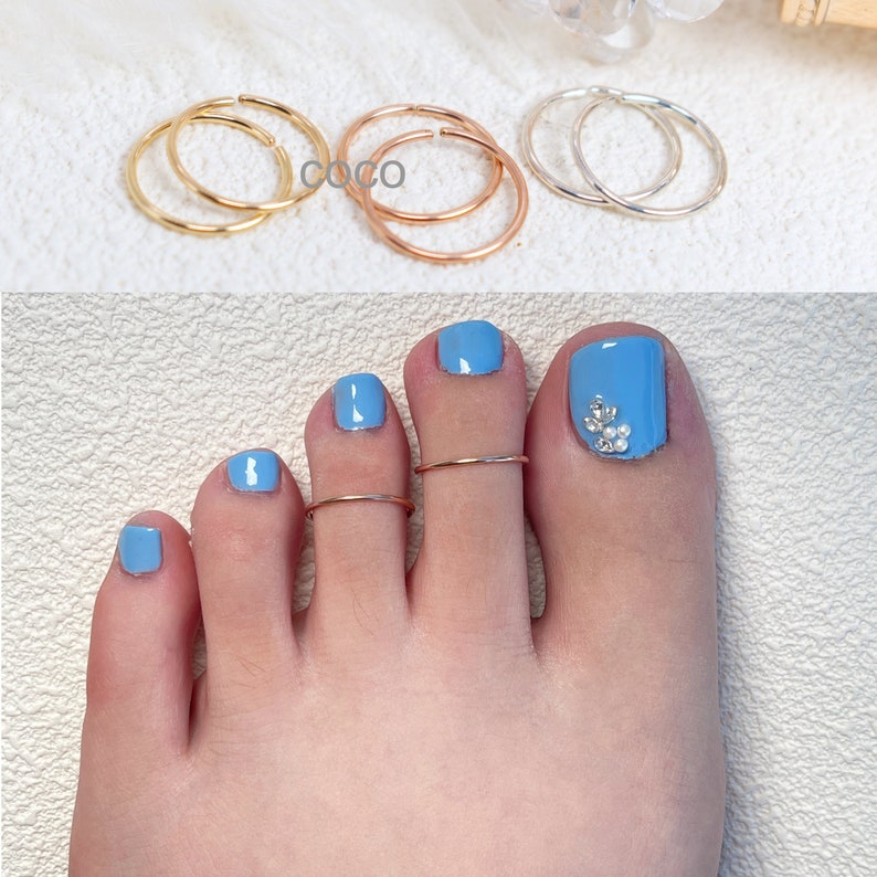 May include: A collection of thin, circular metal rings in gold, rose gold, and silver tones. Two rose gold rings are worn on the second and third toes of a foot with light blue nail polish, featuring small white accents on the big toe nail.