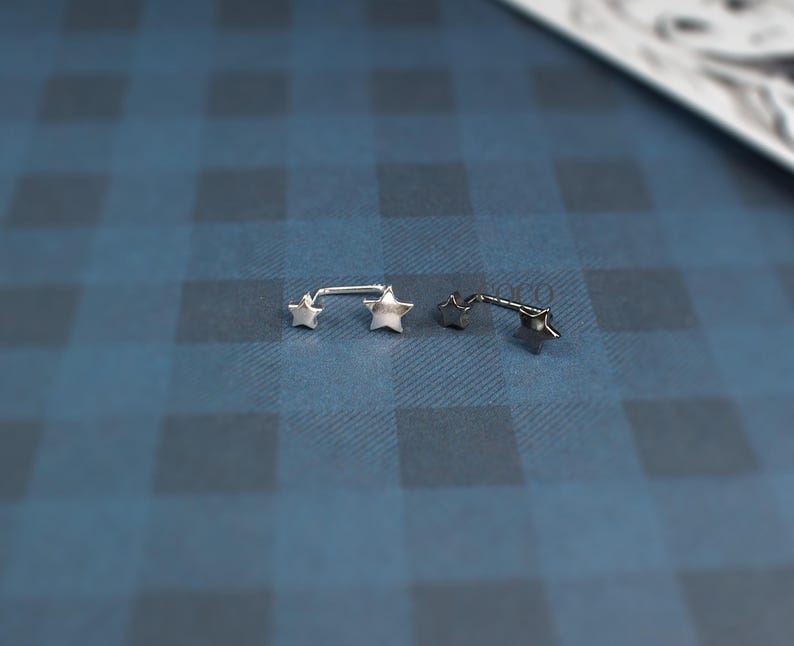 Puede incluir: Dos piercings de barra de metal en tono plateado, cada uno con dos extremos en forma de estrella. Las piezas de joyer&iacute;a est&aacute;n colocadas horizontalmente, mostrando un acabado liso y pulido en los motivos de estrella y las barras rectas.