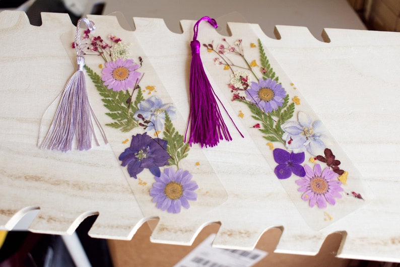 May include: Three handmade bookmarks with pressed flowers and a tassel. The bookmarks are clear and show purple, pink, and white flowers. The tassels are purple and lavender.