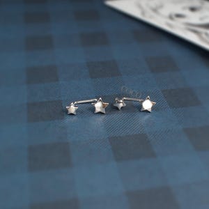 Puede incluir: Un par de pendientes de metal plateado, cada uno con dos estrellas de diferentes tama&ntilde;os conectadas entre s&iacute;. Los pendientes tienen un acabado liso y reflectante, y est&aacute;n colocados horizontalmente para mostrar el dise&ntilde;o.