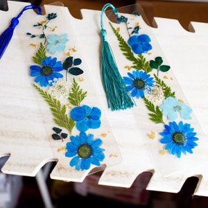 May include: Three blue and white pressed flower bookmarks with blue and teal tassels. The bookmarks are on a white wooden surface.