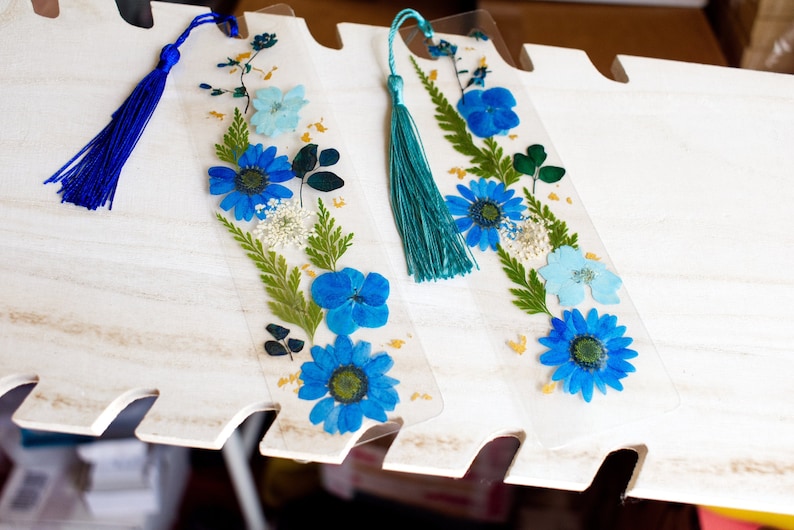 May include: Three blue and white pressed flower bookmarks with blue and teal tassels. The bookmarks are on a white wooden surface.