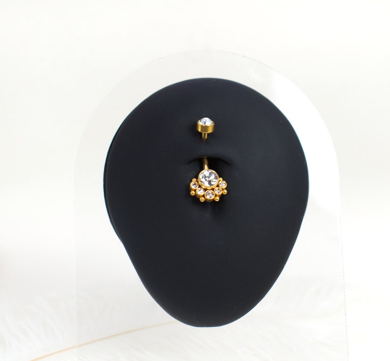 May include: A gold belly button ring with a clear crystal center and smaller crystals surrounding it. The ring is displayed on a black stand.
