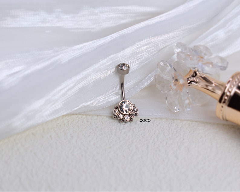May include: A silver belly button ring with a clear crystal center and a row of smaller crystals around it. The ring is on a white fabric background.