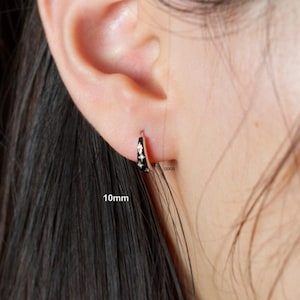 May include: A small silver-toned hoop earring featuring a black enamel inlay with three embedded star-shaped crystals. The earring is labelled with the text 10mm and coco, indicating a diameter of 10 millimetres for this piece of jewellery.