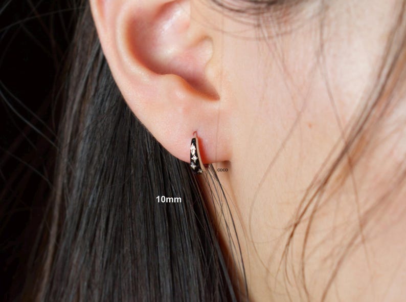 May include: A small silver-toned hoop earring featuring a black enamel inlay with three embedded star-shaped crystals. The earring is labelled with the text 10mm and coco, indicating a diameter of 10 millimetres for this piece of jewellery.