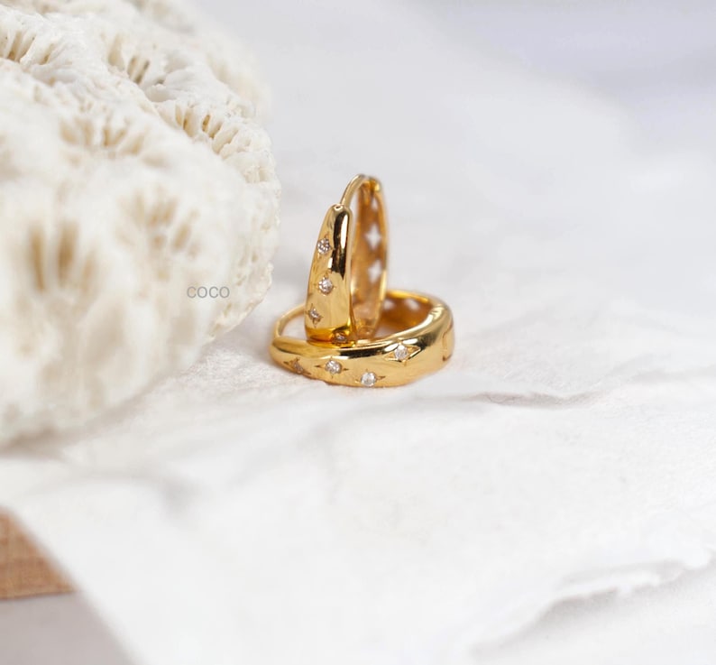 May include: A pair of gold hoop earrings featuring a polished finish and small, star-shaped engravings with embedded clear stones. The earrings are circular with a hinged closure mechanism, displaying a bright metallic lustre and intricate detailing.