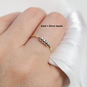 May include: A gold-colored ring with three silver beads. The ring is on a finger, with the text "Gold + Silver beads" visible. The ring is a simple, elegant piece of jewelry.