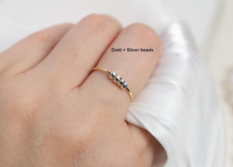 May include: A gold-colored ring with three silver beads. The ring is on a finger, with the text "Gold + Silver beads" visible. The ring is a simple, elegant piece of jewelry.