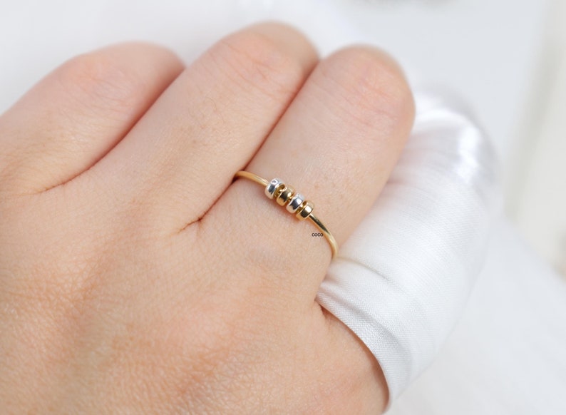 May include: A delicate gold ring with three small, colorful beads. The beads are silver, gold, and gold. The ring is on a finger, with a soft, white background.