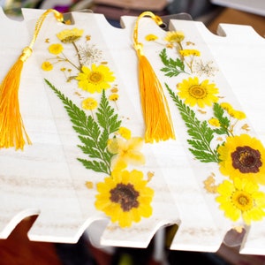 May include: Two yellow pressed flower bookmarks with gold accents and yellow tassels. The bookmarks are on a white wooden surface.