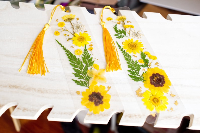 May include: Two yellow pressed flower bookmarks with gold accents and yellow tassels. The bookmarks are on a white wooden surface.