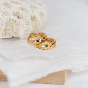 May include: Two gold-toned rings featuring a polished finish and small, embedded clear stones arranged in a star-like pattern. The rings have a smooth, rounded band design and are positioned together, showcasing their reflective metallic surface.