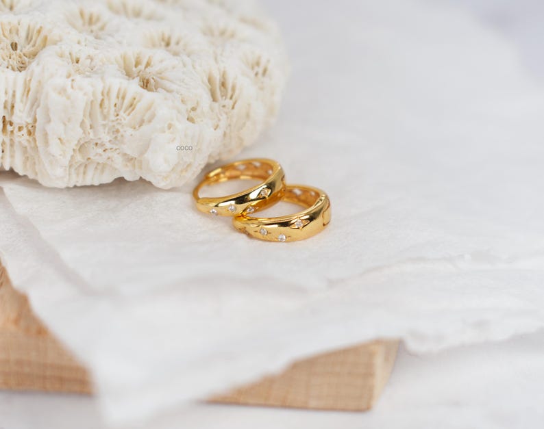 May include: Two gold-toned rings featuring a polished finish and small, embedded clear stones arranged in a star-like pattern. The rings have a smooth, rounded band design and are positioned together, showcasing their reflective metallic surface.