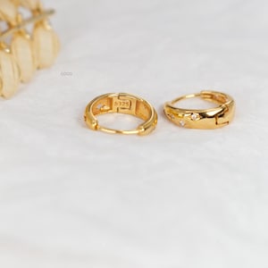 May include: A pair of gold-toned hoop earrings featuring small embedded clear stones and the engraving S925 on the inner band. The earrings have a hinged closure mechanism and a polished finish, with the word coco printed near the top left corner.