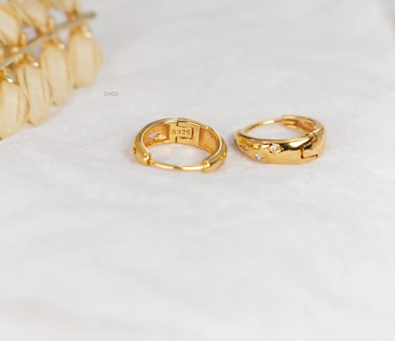 May include: A pair of gold-toned hoop earrings featuring small embedded clear stones and the engraving S925 on the inner band. The earrings have a hinged closure mechanism and a polished finish, with the word coco printed near the top left corner.