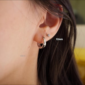 May include: A silver-toned hoop earring featuring small embedded clear stones, positioned in an earlobe. The hoop is labelled with the text 12mm, indicating its size. A small, singular stud earring is also visible in the upper ear piercing.