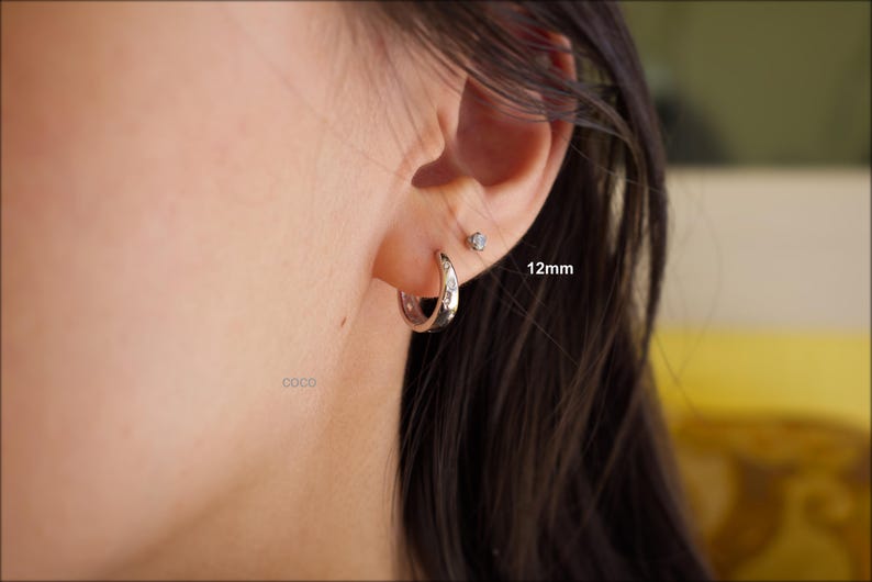 May include: A silver-toned hoop earring featuring small embedded clear stones, positioned in an earlobe. The hoop is labelled with the text 12mm, indicating its size. A small, singular stud earring is also visible in the upper ear piercing.