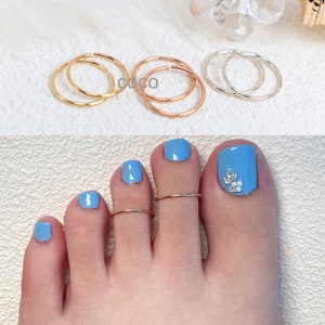 May include: A collection of thin, circular metal rings in gold, rose gold, and silver tones. The rings are shown individually and worn on the second and third toes, which feature light blue nail polish and a small cluster of clear rhinestones.
