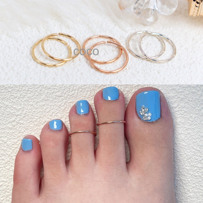 May include: A collection of thin, circular metal rings in gold, rose gold, and silver tones. The rings are shown individually and worn on the second and third toes, which feature light blue nail polish and a small cluster of clear rhinestones.