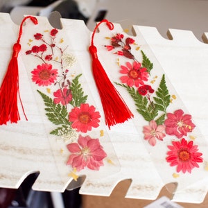 May include: Three clear bookmarks with pressed flowers and red tassels. The bookmarks feature a variety of pink and red flowers, green ferns, and gold accents.