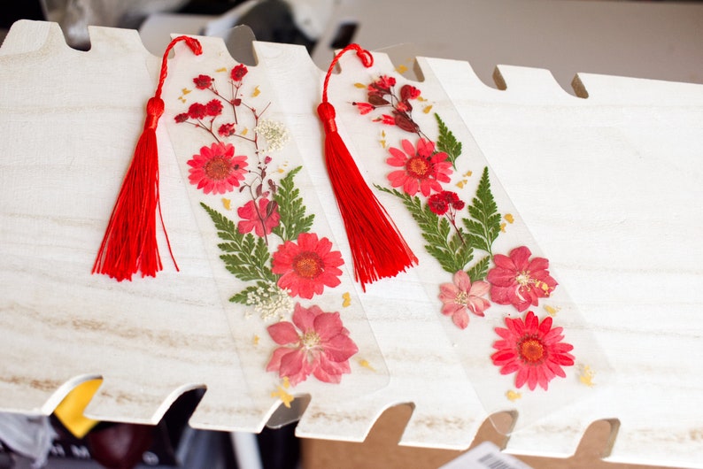 May include: Three clear bookmarks with pressed flowers and red tassels. The bookmarks feature a variety of pink and red flowers, green ferns, and gold accents.