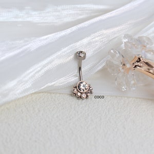 May include: A silver belly button ring with a clear crystal center and smaller crystals surrounding it. The ring is on a white fabric background.