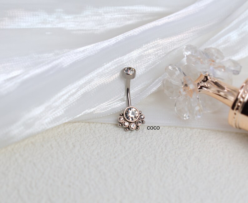 May include: A silver belly button ring with a clear crystal center and smaller crystals surrounding it. The ring is on a white fabric background.