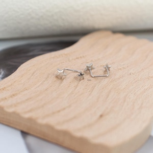 Puede incluir: Dos piercings de oreja de metal plateado, cada uno con un peque&ntilde;o dije en forma de estrella en un extremo y un poste recto. Las piezas de joyer&iacute;a son met&aacute;licas con un acabado pulido, dise&ntilde;adas para el cuerpo y expuestas sobre una superficie.