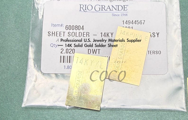 May include: Sheet solder labeled "14KY" and "14K Solid Gold Solder Sheet" from Rio Grande, a U.S. jewelry materials supplier. The image shows two small, rectangular pieces of gold-colored metal, with text and markings.