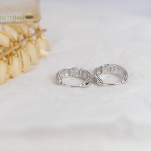 May include: A pair of small silver hoop earrings with a hinged closure mechanism. Each earring is stamped with the text S925 on the inner surface, indicating sterling silver composition. The hoops have a polished metallic finish and a circular shape.