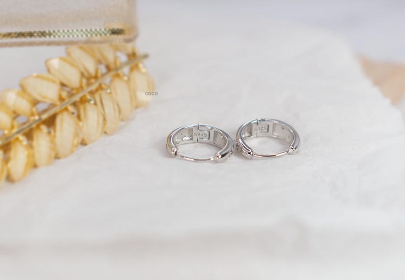 May include: A pair of small silver hoop earrings with a hinged closure mechanism. Each earring is stamped with the text S925 on the inner surface, indicating sterling silver composition. The hoops have a polished metallic finish and a circular shape.