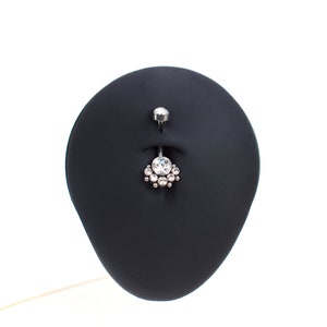 May include: A silver belly button ring with a clear crystal center and smaller crystals surrounding it. The ring is displayed on a black stand.