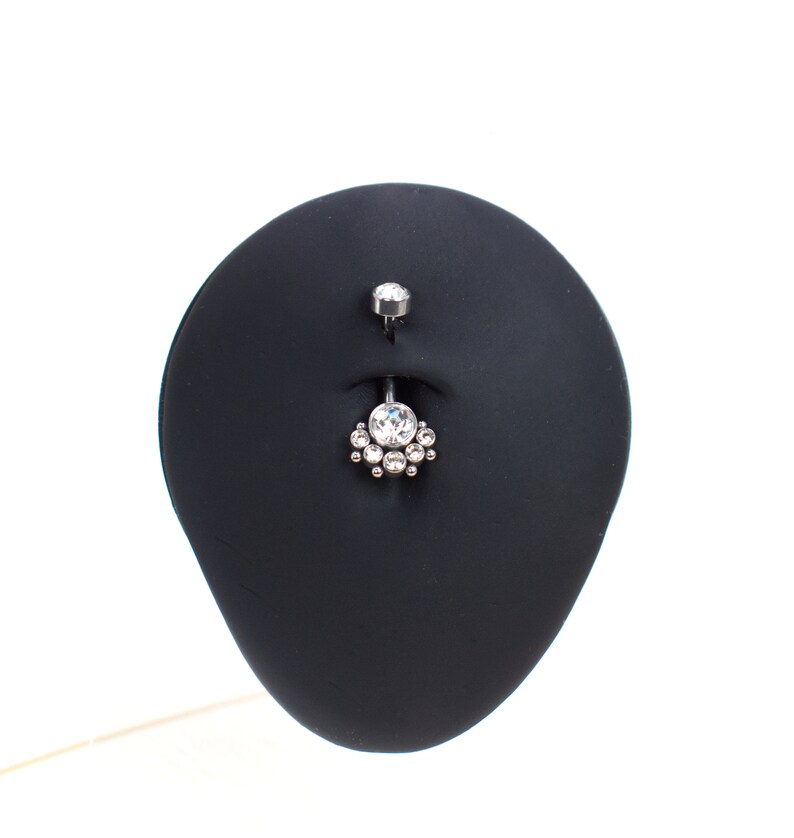 May include: A silver belly button ring with a clear crystal center and smaller crystals surrounding it. The ring is displayed on a black stand.