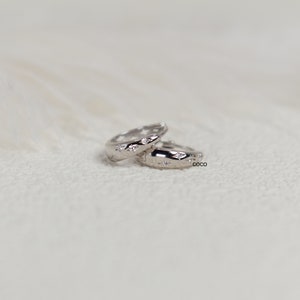 May include: A pair of small silver hoop earrings featuring a polished metallic finish and embedded star-shaped accents. Each hoop is engraved with the word coco on the side, showcasing a clean, reflective surface and a circular design.