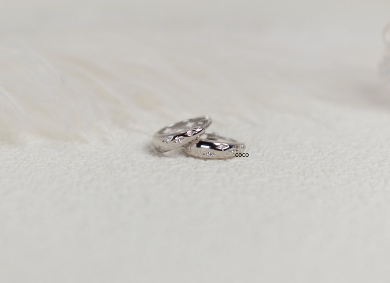 May include: A pair of small silver hoop earrings featuring a polished metallic finish and embedded star-shaped accents. Each hoop is engraved with the word coco on the side, showcasing a clean, reflective surface and a circular design.