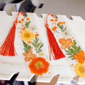 May include: Two handmade bookmarks with pressed flowers and red tassels. The bookmarks feature a variety of dried flowers, including yellow, orange, and white flowers, and green leaves.