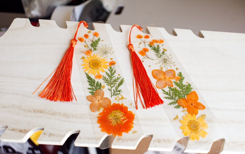 May include: Two handmade bookmarks with pressed flowers and red tassels. The bookmarks feature a variety of dried flowers, including yellow, orange, and white flowers, and green leaves.